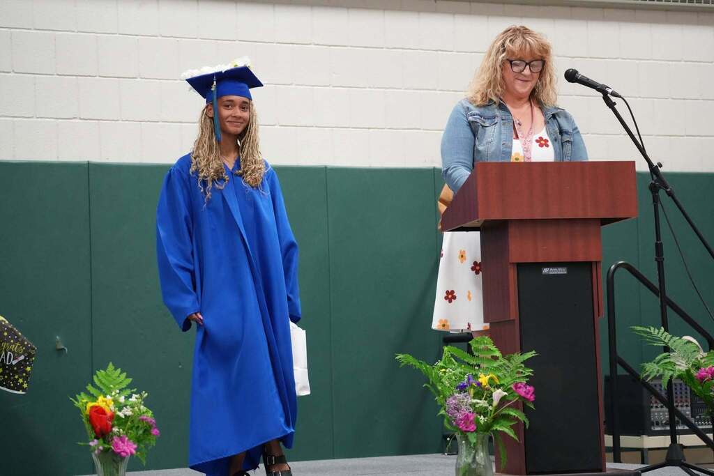Graduate wearing a decorated graduation cap stands beside a staff member speaking at the podium during the Turning Point graduation ceremony.
