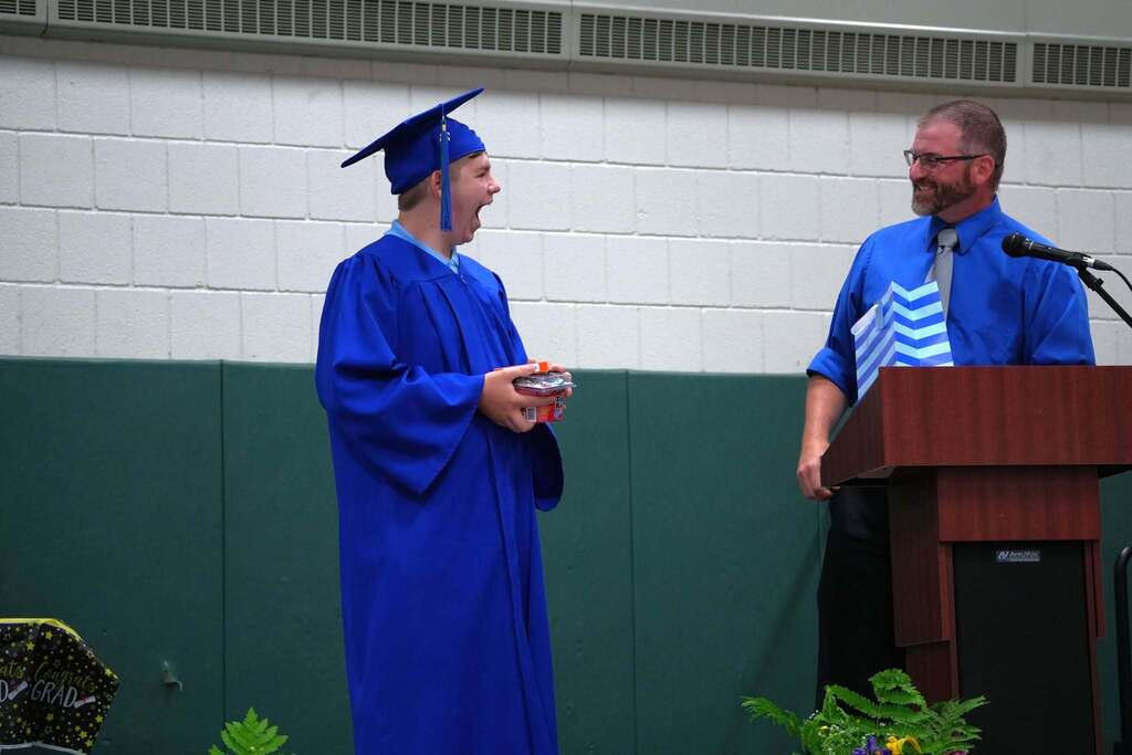 Graduate in a blue cap and gown smiles with surprise while accepting a gift during the Turning Point graduation ceremony as a staff member looks on from the podium.