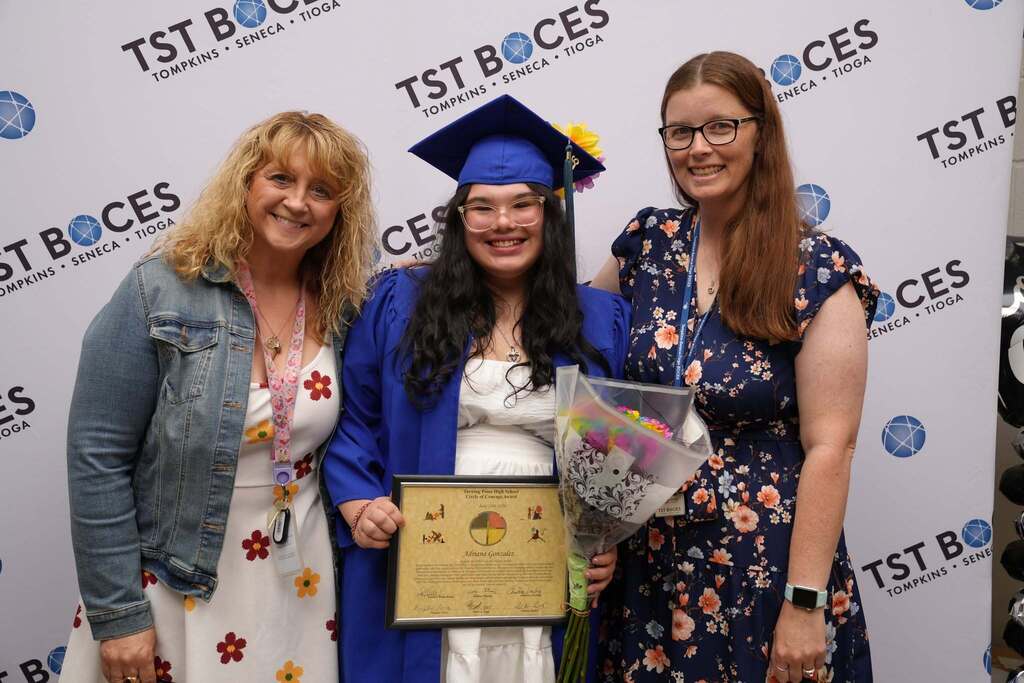 Graduate holding a Circle of Courage Award certificate and bouquet poses with two staff members in front of a TST BOCES backdrop.