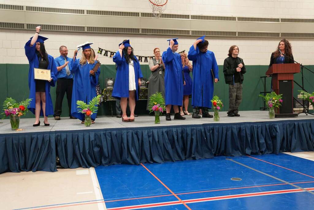 Turning Point graduates stand on stage in blue caps and gowns during the tassel ceremony while family members and staff applaud.