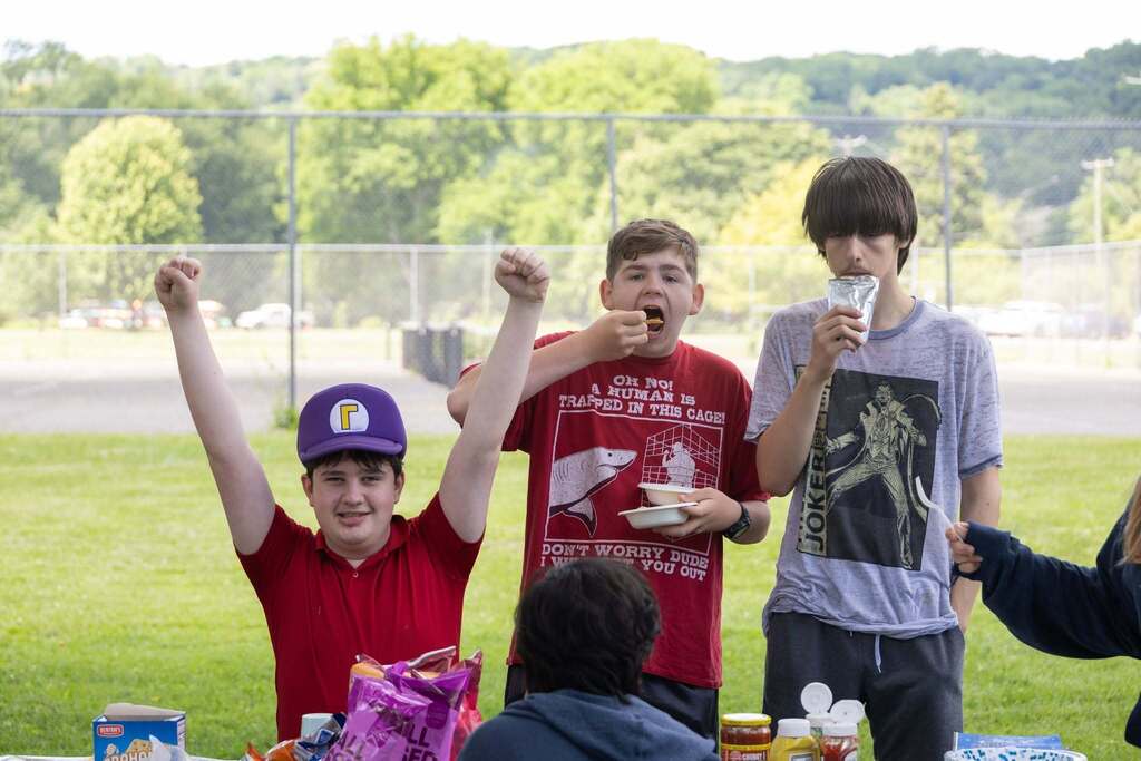 Student raises both arms in celebration while standing at a picnic table with classmates enjoying snacks and lunch during the Turning Point end-of-year BBQ at Stewart Park.