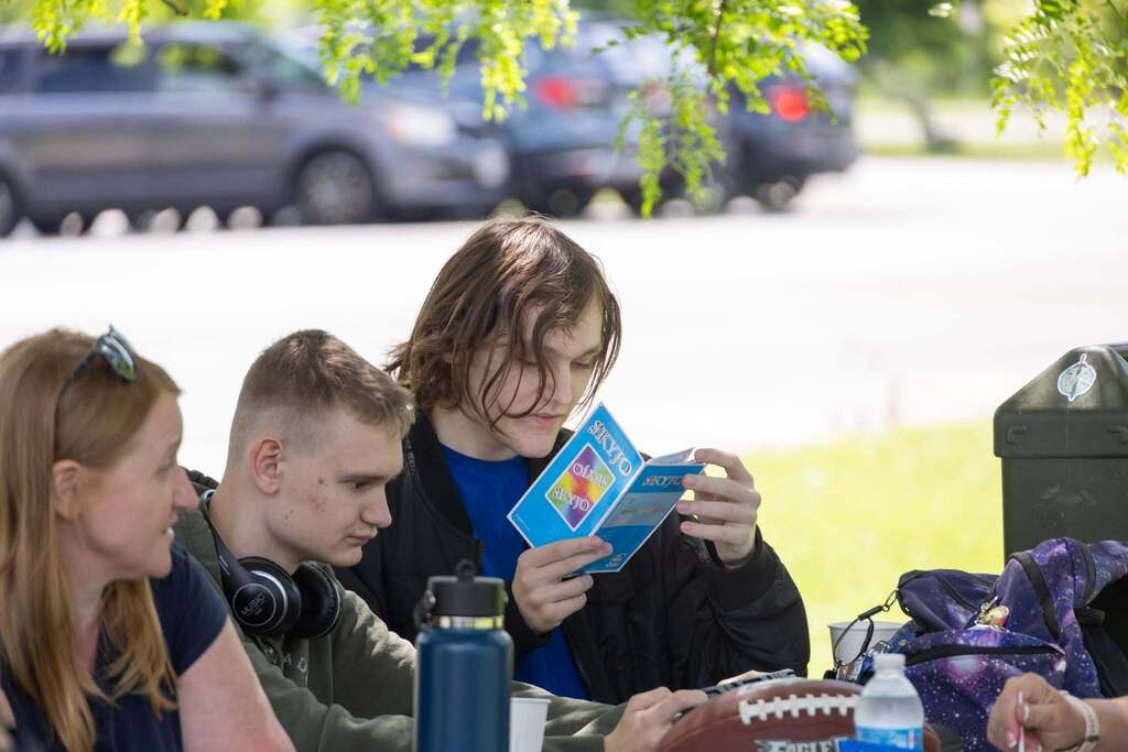 Students sit together at a picnic table, reading game cards and spending time together during the end-of-year celebration.