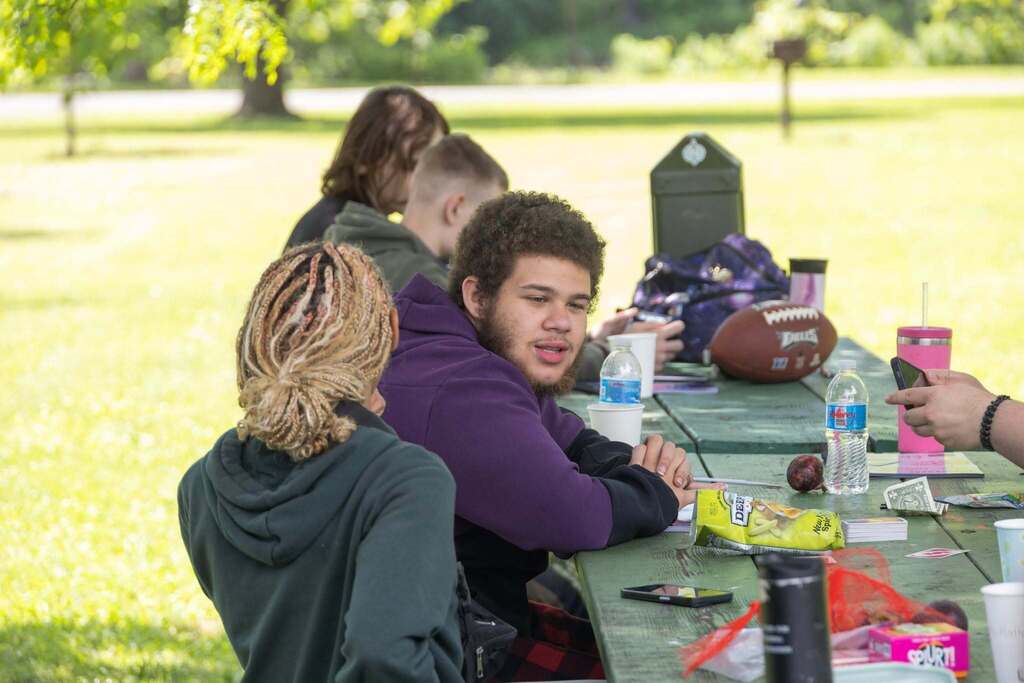 Students sit together at a picnic table talking, eating snacks, and enjoying the outdoor BBQ celebration.