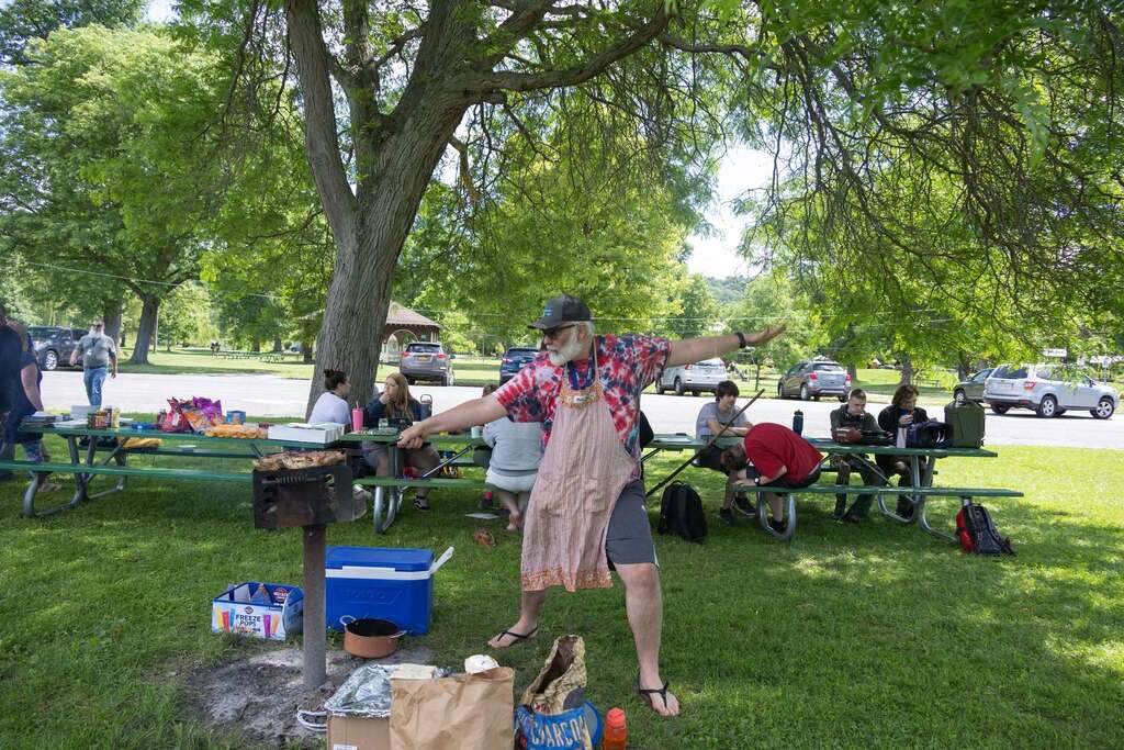 Staff member grills food and playfully poses with arms outstretched while students gather at picnic tables in the background.