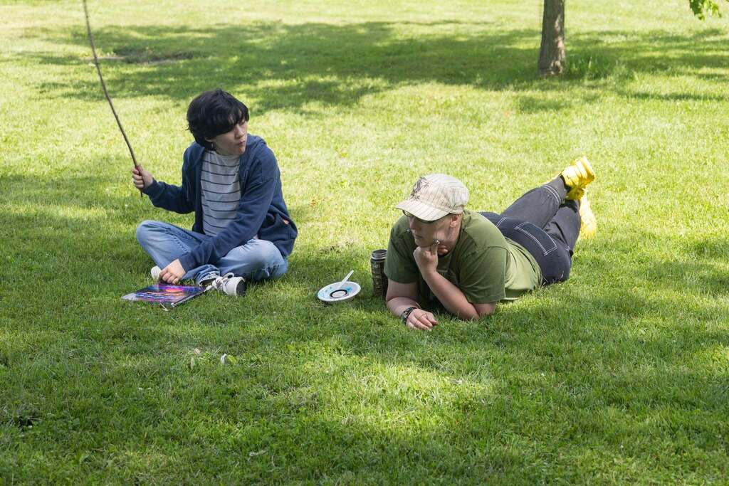 Student and staff member relax on the grass in the shade, talking and enjoying the outdoor celebration.