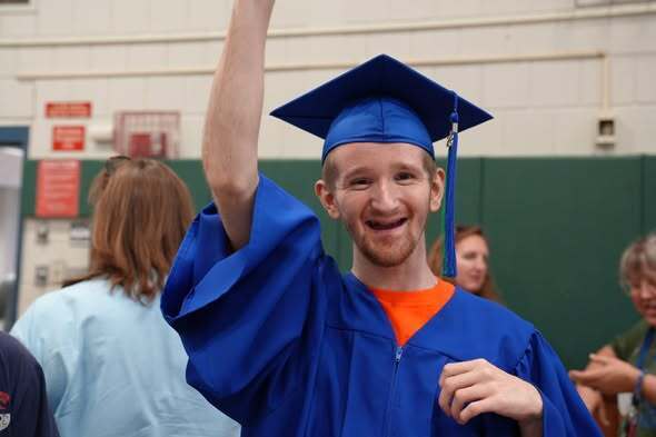 Graduate in a blue cap and gown smiles and raises a hand in celebration during the Bridges & Steps graduation ceremony.