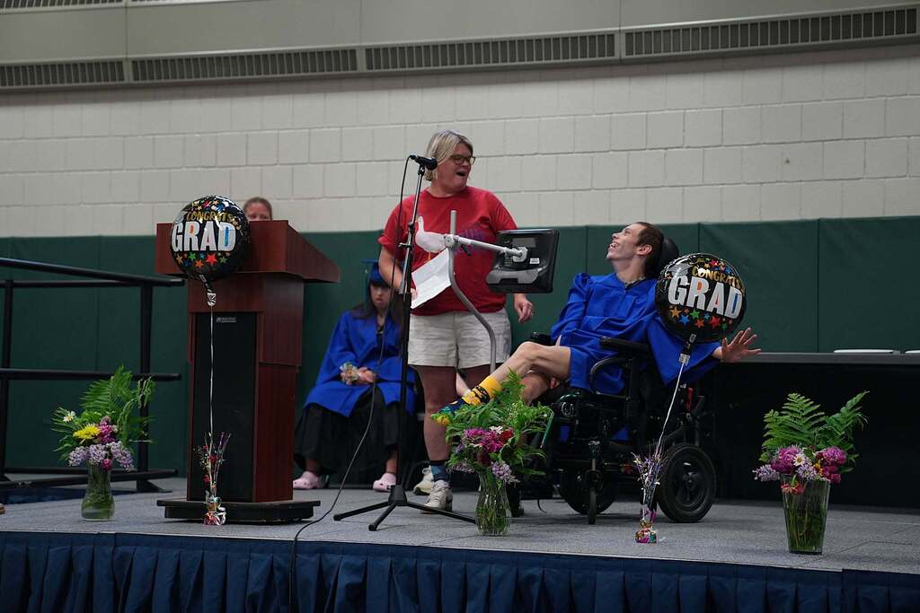 Staff member reads remarks at the microphone while a graduate in a wheelchair smiles beside them on stage during the ceremony.