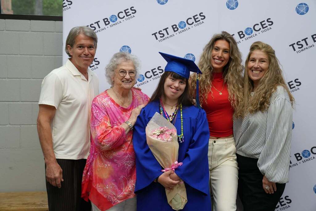Graduate and family members share joyful smiles while posing with a bouquet of flowers in front of a TST BOCES backdrop.