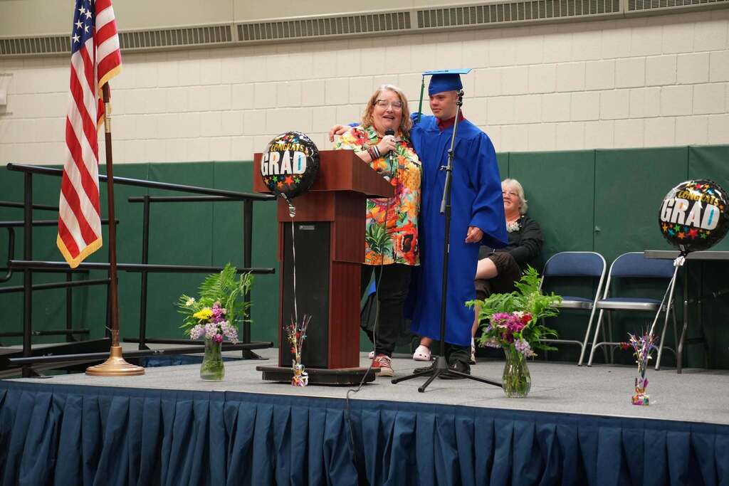 Graduate stands beside a staff member at the podium during the graduation ceremony while speaking to the audience.