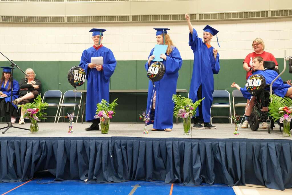 Bridges & Steps graduates in blue caps and gowns stand on stage during their graduation ceremony. One graduate speaks while holding notes, another holds a folder, and a graduate raises a finger in celebration. A graduate in a wheelchair smiles alongside staff members as colorful flower arrangements and graduation balloons decorate the stage.