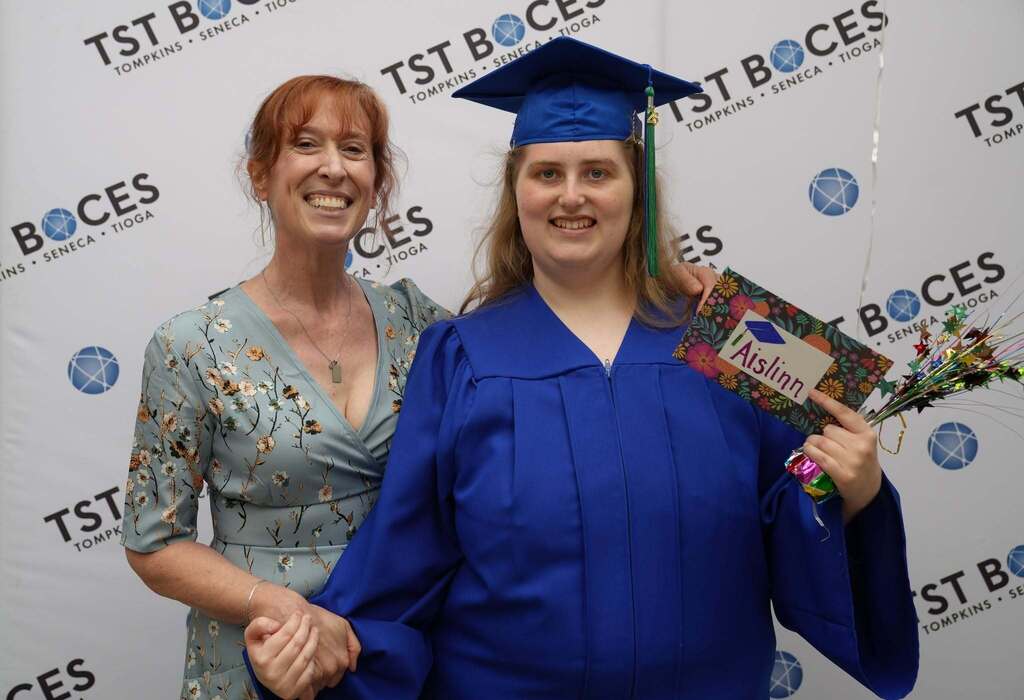 Graduate in a blue cap and gown poses with a family member while holding a decorated graduation cap and celebration gift.