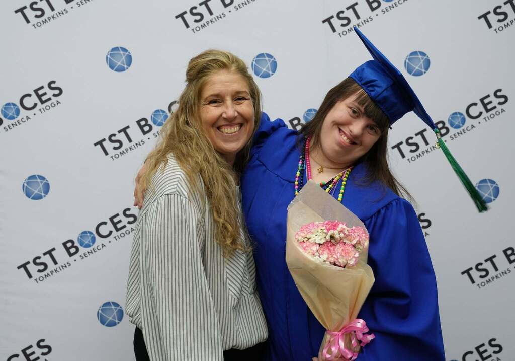 Graduate holding a bouquet of pink flowers poses with a smiling family member in front of a TST BOCES graduation backdrop.