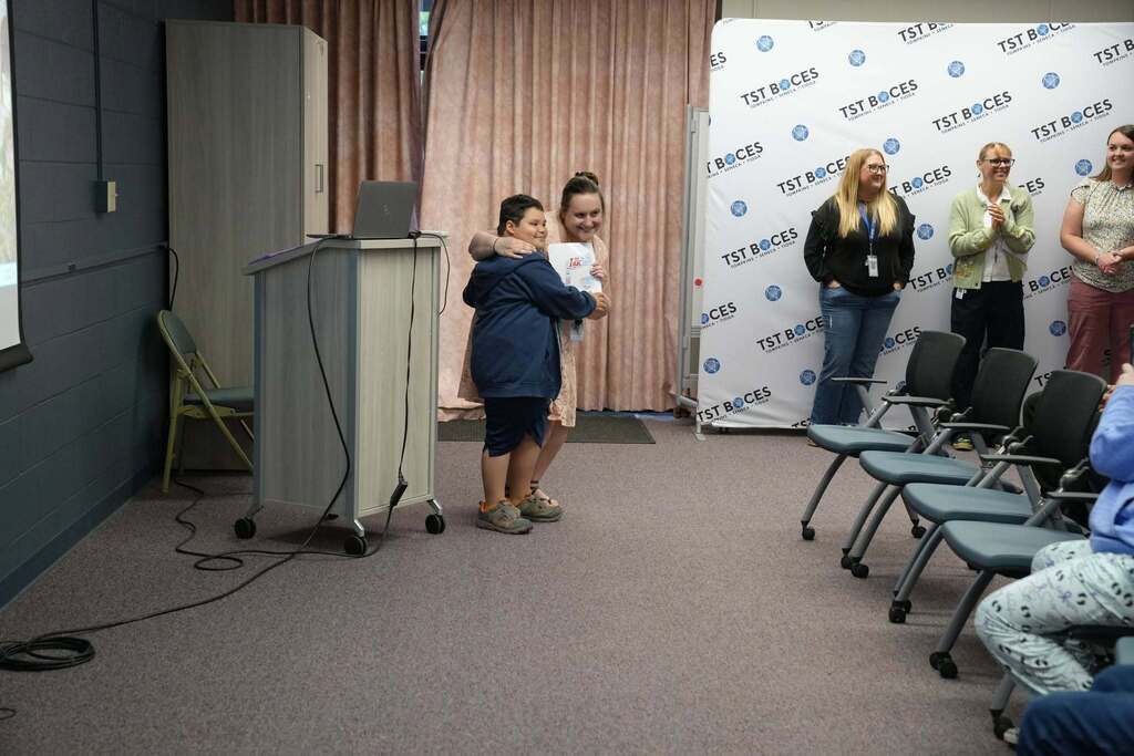 A student receives a certificate and a congratulatory hug from a staff member during the Turning Point Elementary end-of-year awards ceremony as staff and families applaud nearby