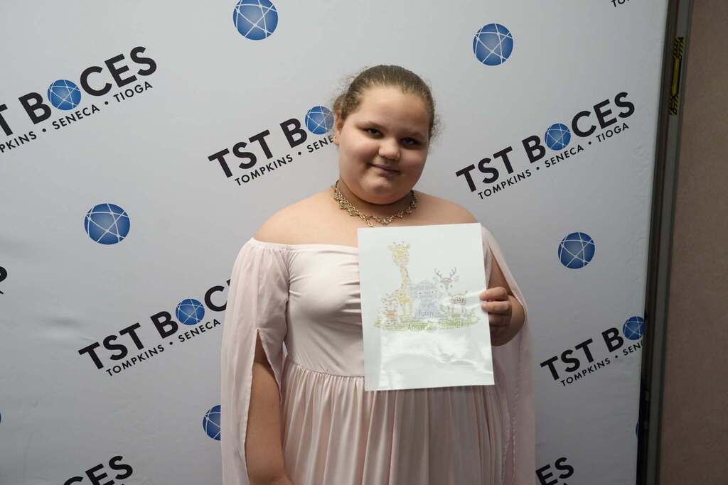 A student smiles while holding a personalized word art print featuring two giraffes made from descriptive words, posing in front of a TST BOCES backdrop.