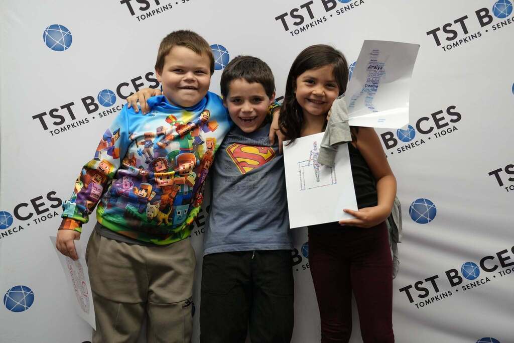 Three Turning Point Elementary students smile with their arms around each other while proudly displaying their personalized word art prints in front of a TST BOCES backdrop.