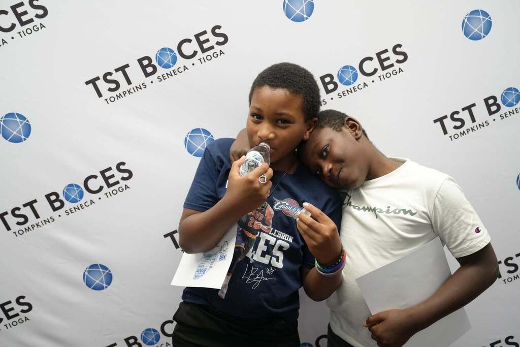Two Turning Point Elementary students lean against each other and smile while posing in front of a TST BOCES backdrop, each holding papers from the celebration.