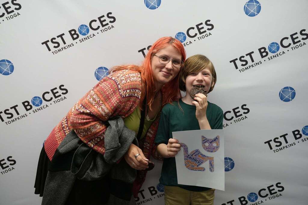 A student and family member pose together in front of a TST BOCES backdrop while the student smiles and holds a personalized word art print and a cookie.