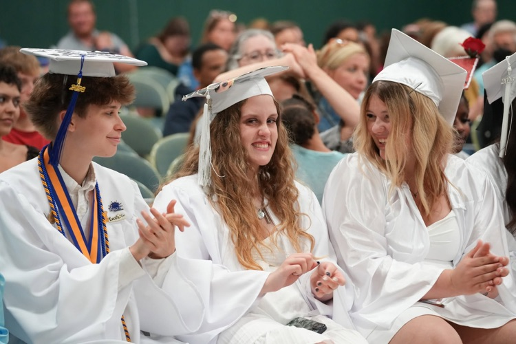 Three graduates in white caps and gowns sit together before the ceremony, smiling, clapping, and sharing a laugh with one another as family members watch from the audience behind them.