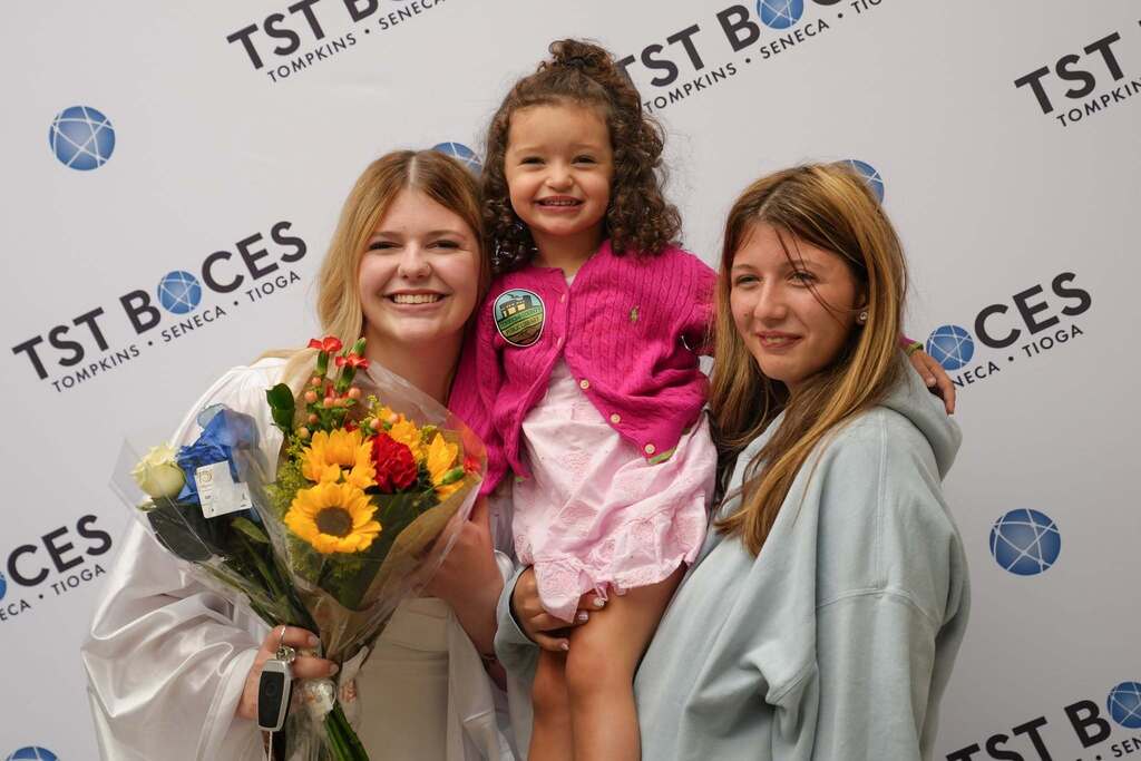 A 2026 graduate in a white cap and gown smiles while posing with a young child and another family member in front of a TST BOCES backdrop, holding a bouquet of flowers after the ceremony.