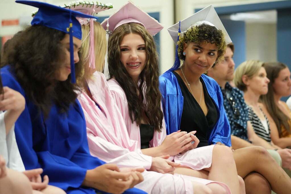 Four graduates in blue, pink, and white caps and gowns sit together smiling and talking while waiting for the graduation ceremony to begin.
