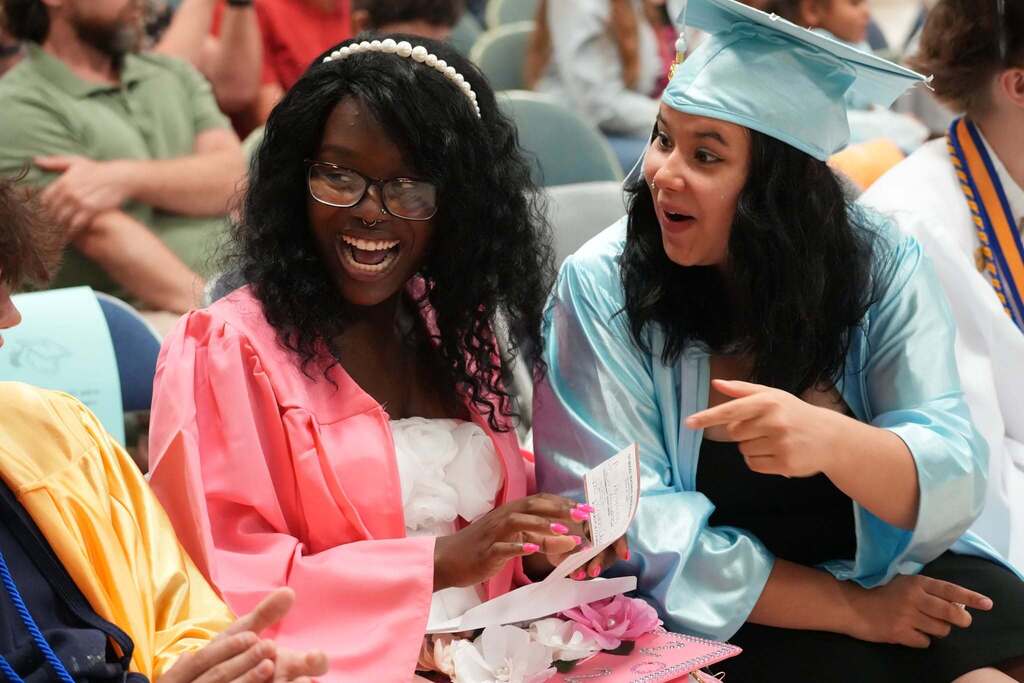 Two graduates in pink and light blue caps and gowns laugh together while looking at a note one of them is holding before the graduation ceremony begins.