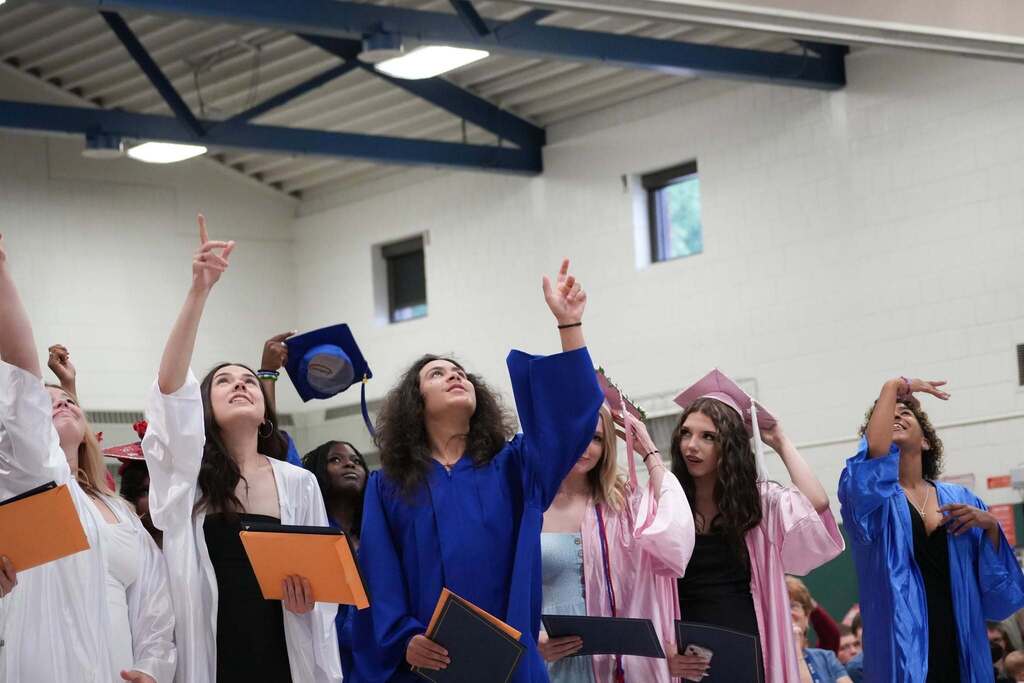 Graduates in blue, white, and pink caps and gowns point toward the ceiling as they watch decorated graduation caps being tossed into the air after the ceremony, while holding their diplomas.