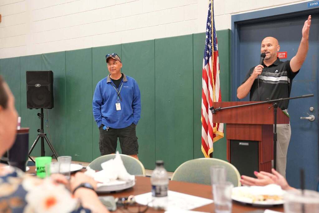 A TST BOCES administrator speaks at the podium during the Annual Hot Dog Day celebration while another staff member stands nearby. Employees seated at tables listen during the program.
