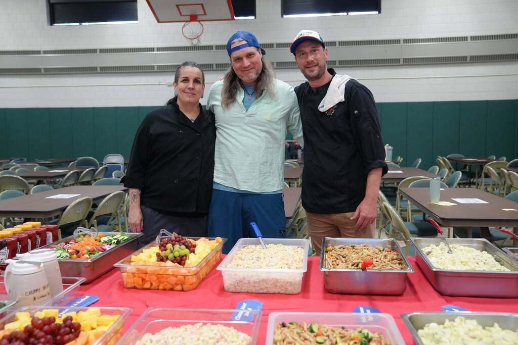 Three members of the TST BOCES Food Service team stand behind a buffet filled with salads, fruit, and side dishes prepared for the Annual Hot Dog Day celebration.