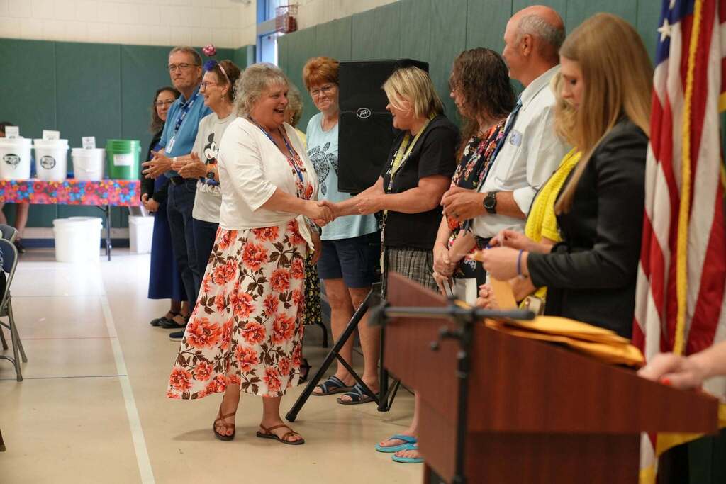 A TST BOCES employee smiles as she shakes hands with a colleague while being recognized for a milestone year of service during the Annual Hot Dog Day celebration. Staff members line the front of the room as attendees applaud.