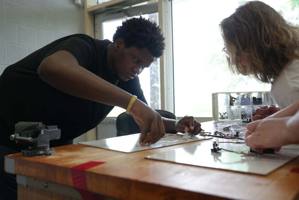 A P-TECH student and teacher work together assembling an electronics project at a classroom workbench, concentrating on placing small components onto a circuit board.