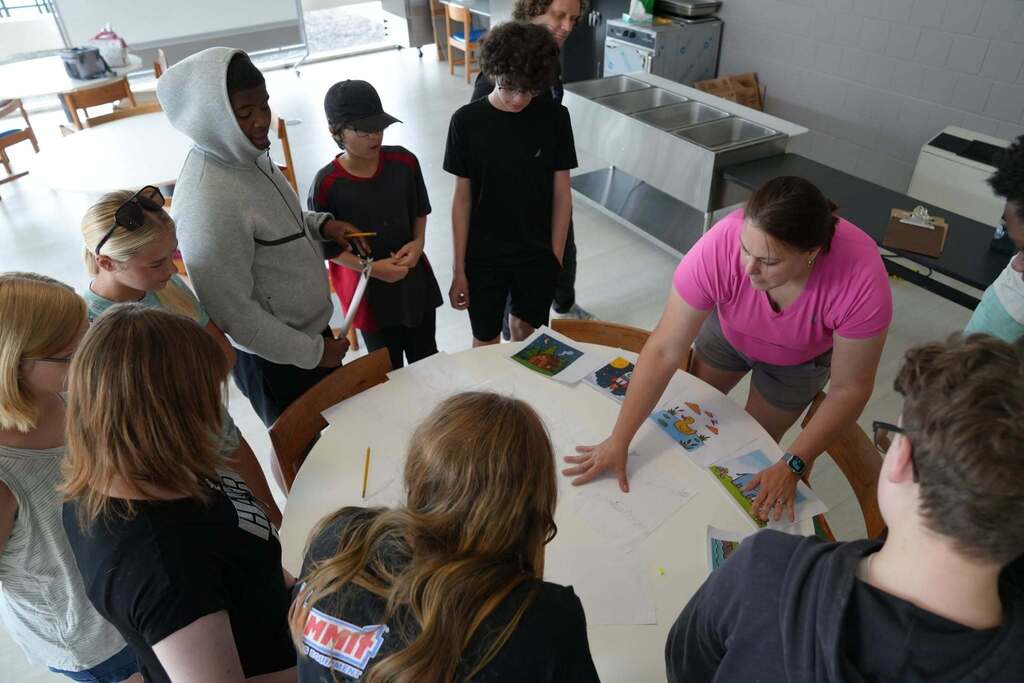 A P-TECH teacher leads a group of incoming students as they collaborate around a table, planning a large team project with sketches and reference images.