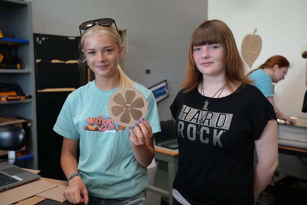 Two incoming P-TECH students smile while holding a laser-cut wooden logo with a flower they created during a hands-on Bridge Week activity.