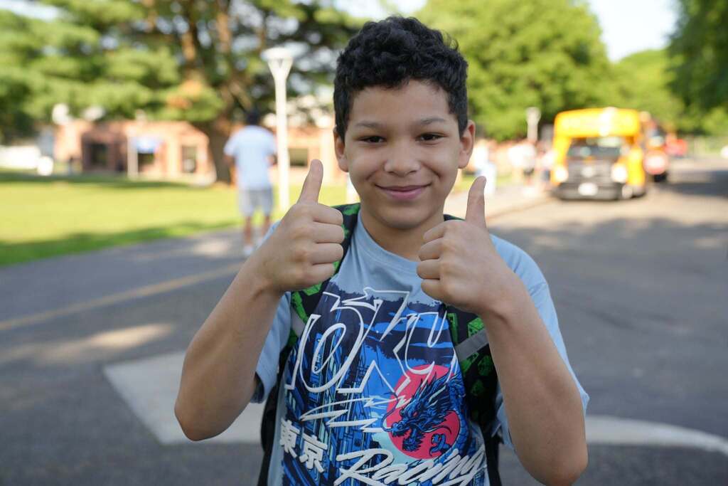 A smiling student wearing a backpack gives two thumbs up while standing outside near school buses during Summer School arrival.