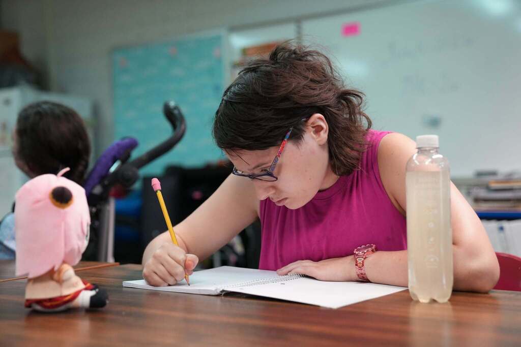 A student concentrates while writing in a notebook at a classroom table during a Summer School lesson. A water bottle and plush toy sit nearby.