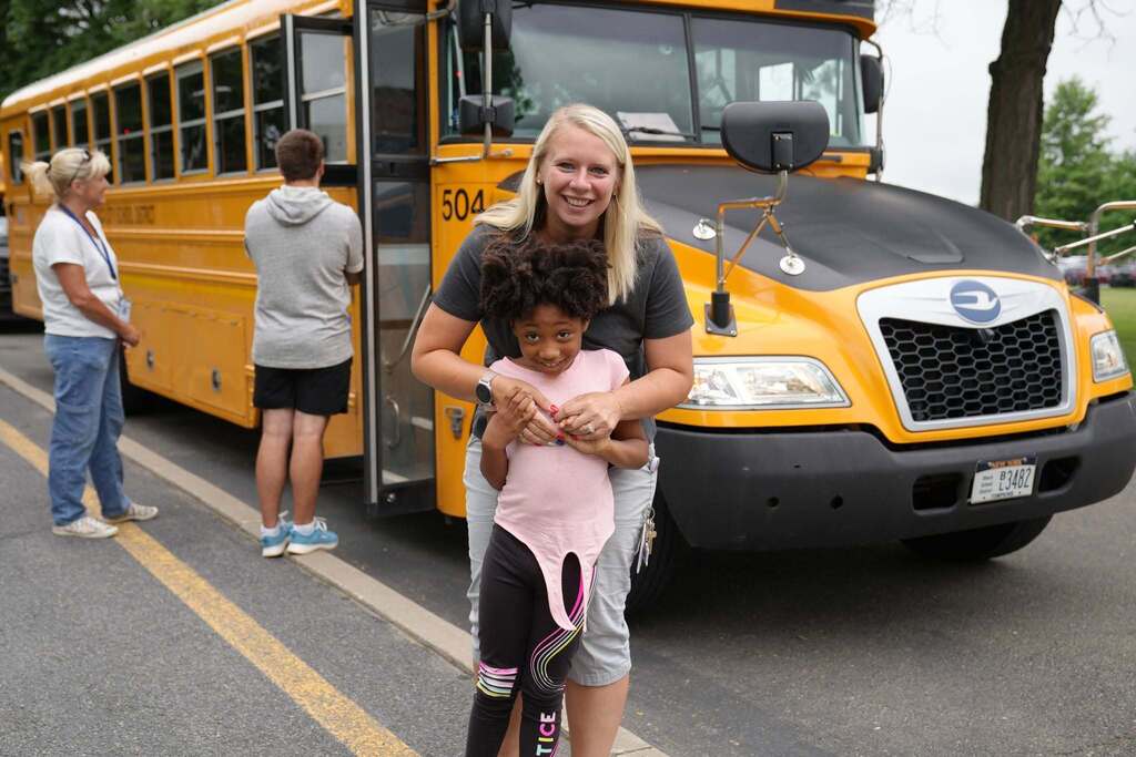 A staff member hugs a smiling young student in front of a parked school bus as students arrive for Summer School.