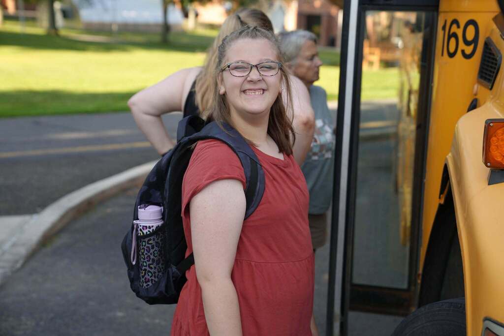 A smiling student wearing a backpack stands beside a school bus before boarding on the first day of Summer School.