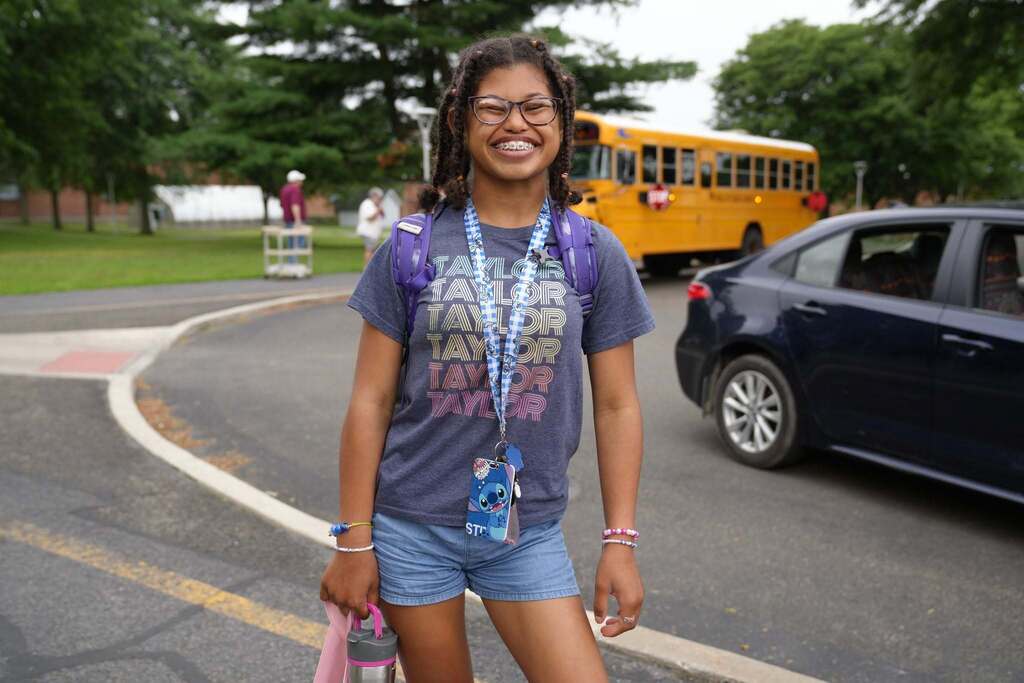A smiling student wearing a backpack and lanyard stands outside near a school bus before heading into Summer School.