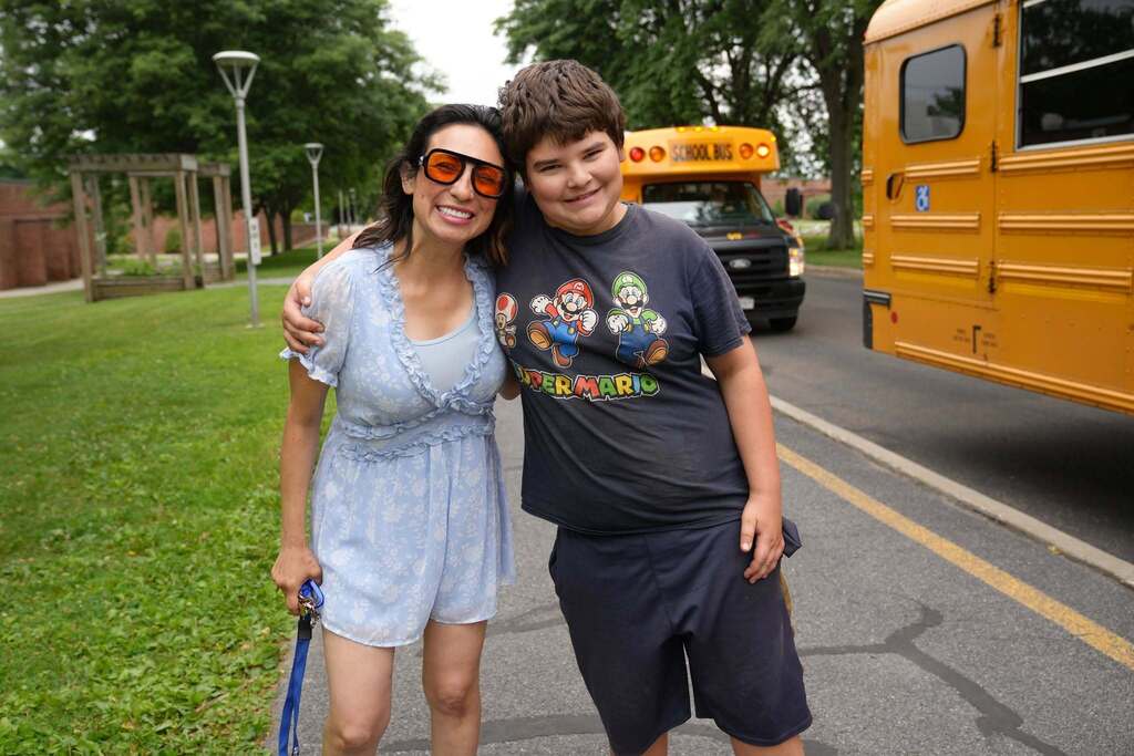A smiling student and staff member stand arm in arm outside beside school buses on the first day of Summer School.