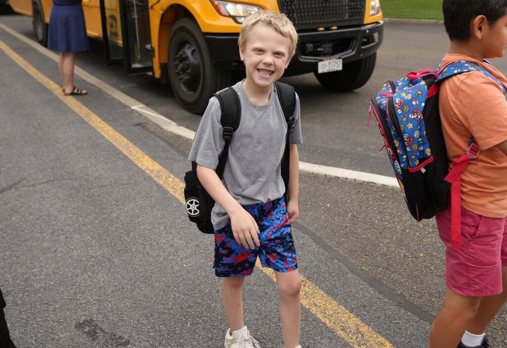 A smiling elementary-aged student wearing a backpack walks toward the school after getting off the bus, while another student walks beside him.