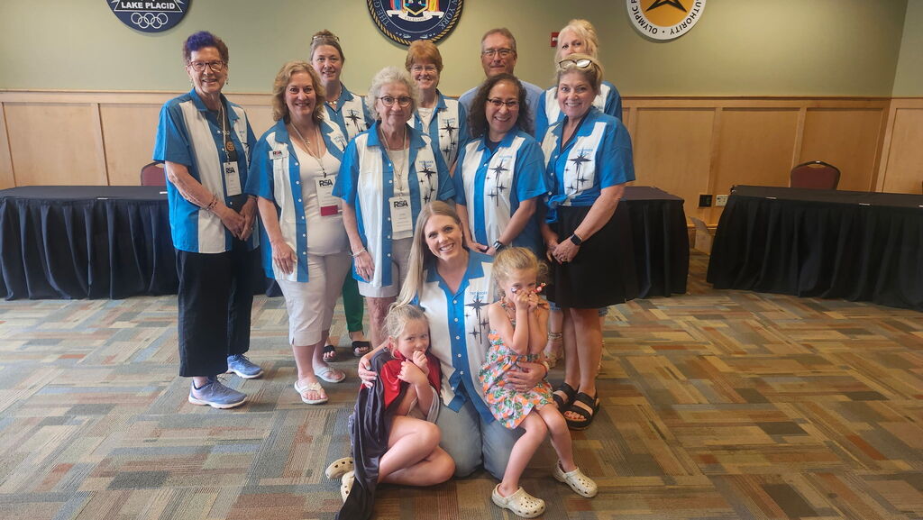 A group of TST BOCES leaders, Board of Education members, and family members pose together inside the conference venue during the 2026 Rural Schools Association of New York State Summer Conference in Lake Placid. The group is wearing matching blue-and-white bowling-style shirts featuring a custom design created by TST BOCES Career & Tech Digital Media students. Two young children sit in front with one attendee, while conference and Olympic-themed signs are displayed on the wall behind the group.
