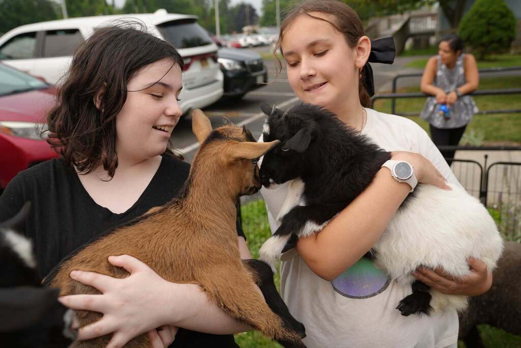 Two students smile while holding baby goats as the curious animals touch noses. Other visitors and animal pens can be seen in the background.