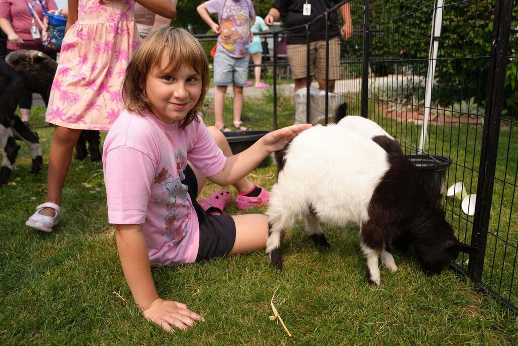 A student sits on the grass and pets a small goat while smiling at the camera during Fall Down Farm's hands-on visit to TST BOCES Summer School.