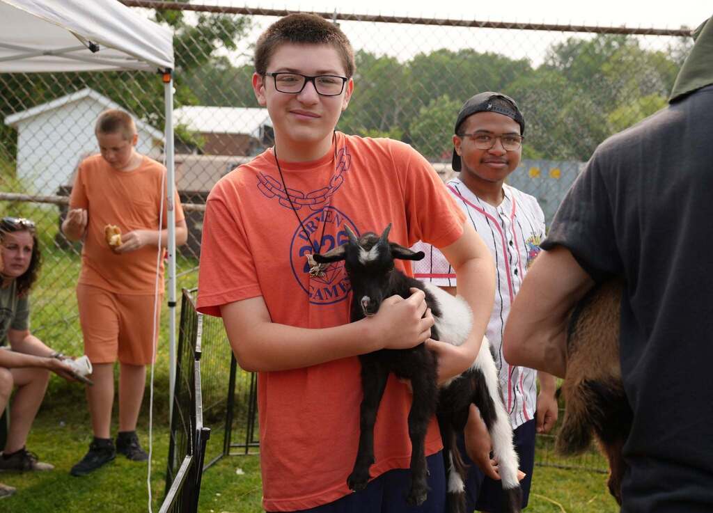 A smiling student holds a baby goat while standing inside the animal enclosure as classmates and staff enjoy the farm visit in the background.