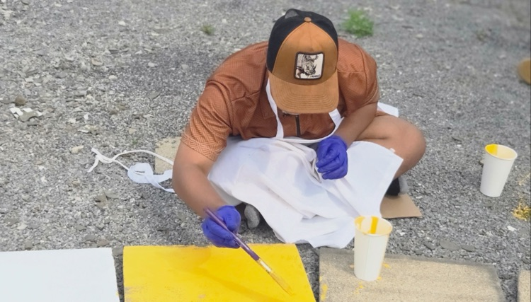 A student wearing a white apron and blue gloves sits outdoors painting a concrete paver bright yellow for the Circle of Courage labyrinth in the Smith School garden. Paint supplies and additional pavers are visible nearby.