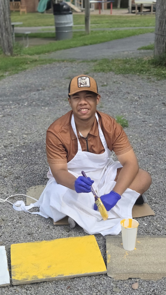 A student wearing a white apron and blue gloves sits outdoors painting a concrete paver bright yellow for the Circle of Courage labyrinth in the Smith School garden smiling at the camera