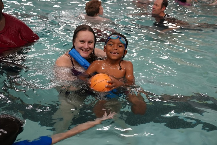 A smiling student wearing swim goggles holds an orange pool basketball while a staff member supports them in the water during a summer school swim trip at Tompkins Cortland Community College.