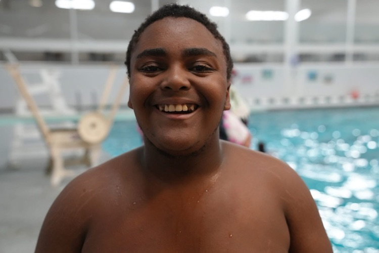 A smiling student stands near the edge of the pool with the swimming area blurred in the background.