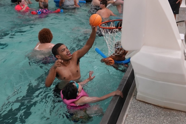 A staff member reaches toward a poolside basketball hoop while students enjoy a game of water basketball during a summer school swim trip.