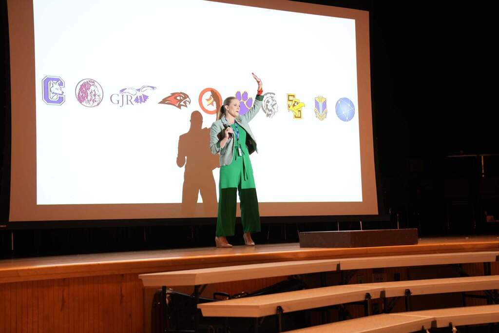Lily Talcott welcomes conference attendees from the auditorium stage with district logos displayed on the screen behind her.