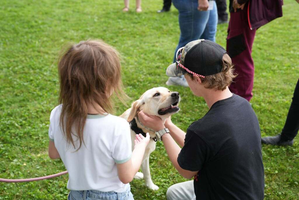 A student kneels on the grass while petting K-9 Meekah, a yellow Labrador retriever from Cortland University Police, as another student reaches out to greet the dog during a campus K-9 demonstration.